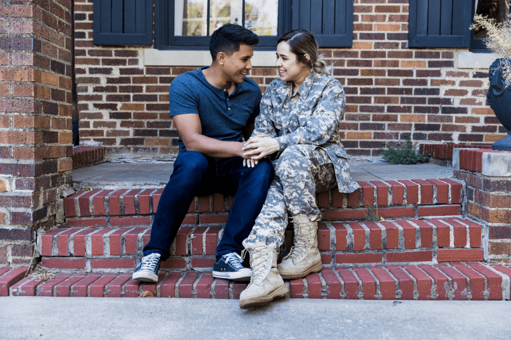 A veteran and her partner sit together on the red brick steps of their house, holding hands. The veteran is wearing Army camo and combat boots, and her partner is dressed casually.