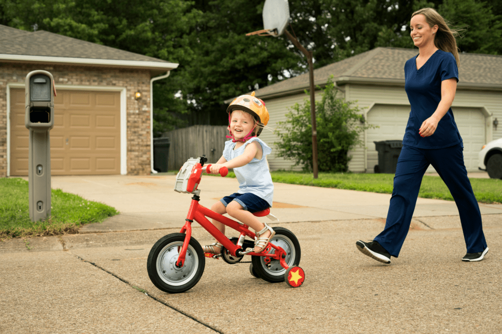 A mother in scrubs walks behind her child, who is riding a red bike with training wheels on a quiet, pretty suburban road.