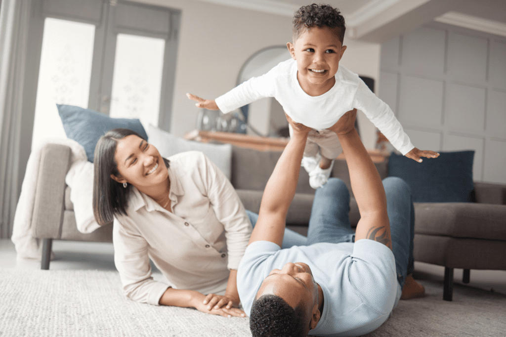 A smiling couple lie on the white carpet of their new house together as the father lifts their baby in the air like an airplane. The baby is smiling and looking around the room.